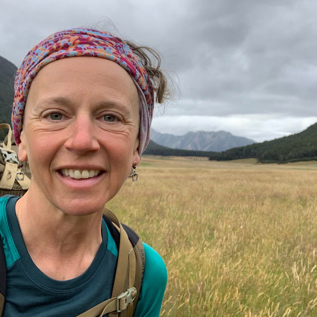 A happy and somewhat blissful hiker leaving early from the crowded hut and walking alone through grassy plains framed by mountains. 