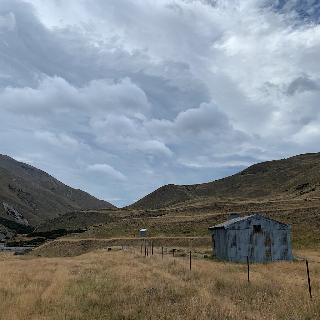 As if out of an old western, Comyns Hut appears along with its long drop in the distance as the clouds build. 